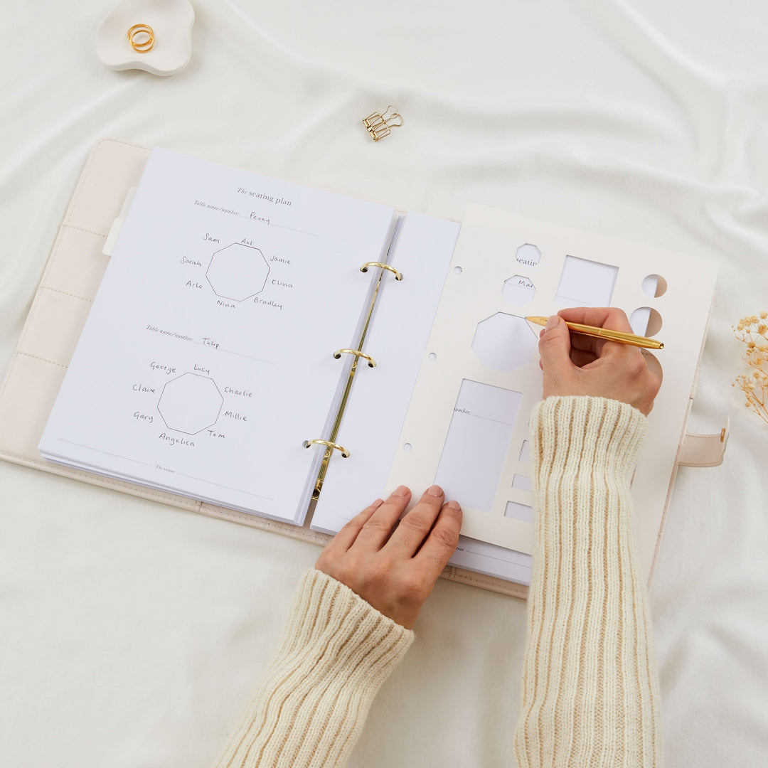 Hands using a table stencil and gold pen to draw a seating plan in a wedding planner binder with completed octagonal table diagrams visible