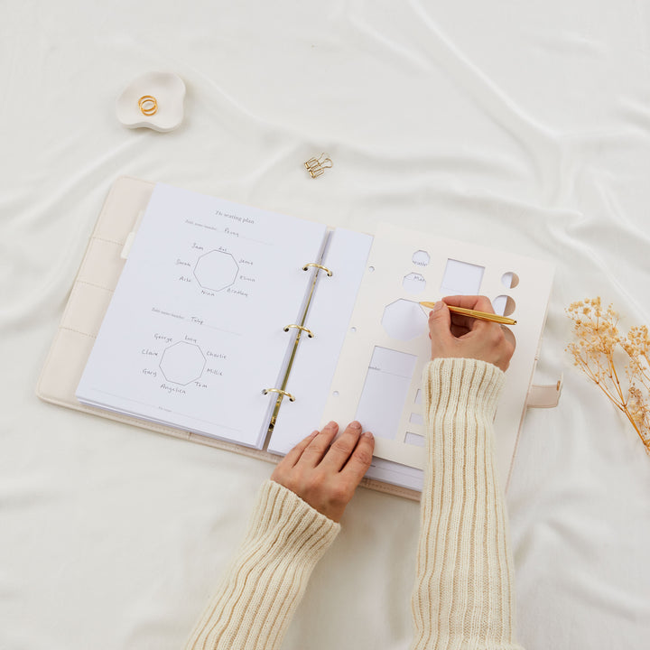 Hands writing guest names onto octagonal table diagrams in a wedding planner binder seating plan page with a gold pen
