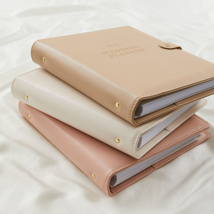 Three wedding planner binders stacked in tan, cream and blush pink vegan leather with gold stud detailing and gold foil cover text