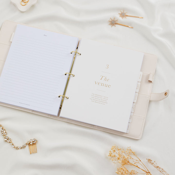 Open wedding planner binder showing a blank notes page alongside the venue section divider with gold foil text on cream vegan leather