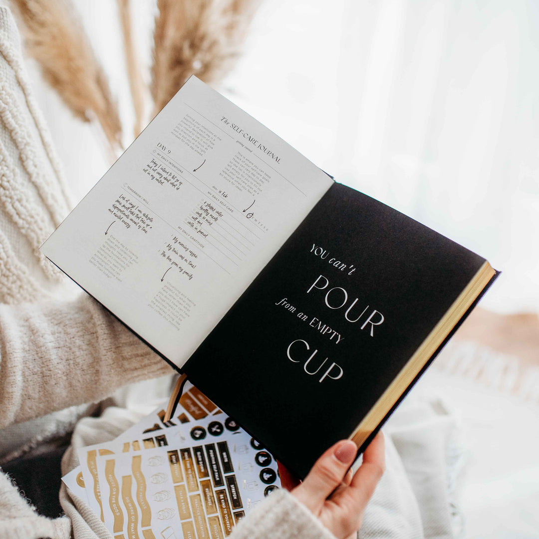 lack journal open to a page reading "You can't pour from an empty cup", with gold edged pages and sticker sheets on a knit blanket