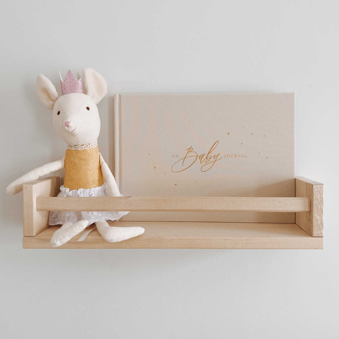 Baby memory book with an ivory cover and gold accents sitting on a baby nursery shelf, surrounded by a soft toy.