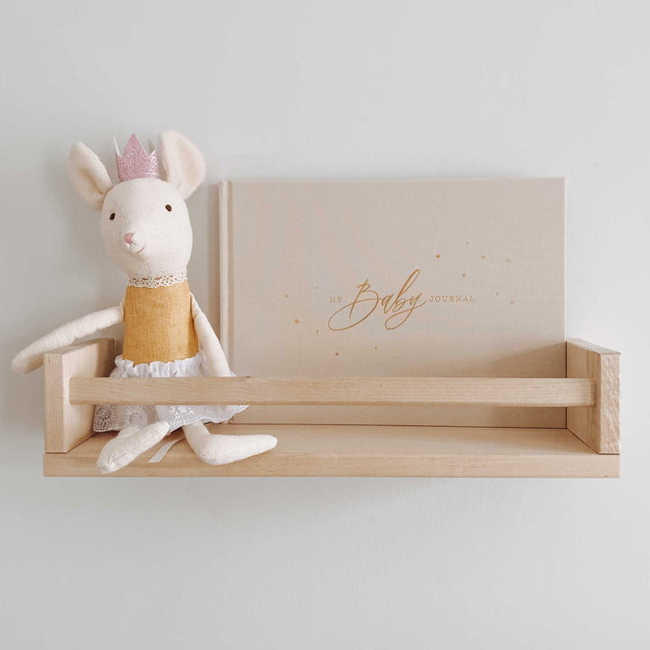 Baby memory book with an ivory cover and gold accents sitting on a baby nursery shelf, surrounded by a soft toy.
