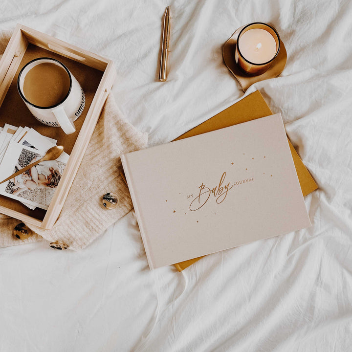 Cover of a baby memory book with an elegant ivory background and gold accents, featuring delicate illustrations of stars, hearts, and a central title that reads 'Baby Journal' in stylish gold lettering.